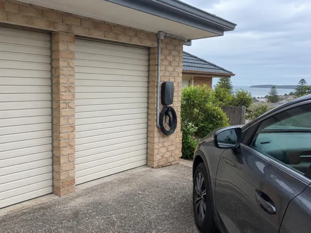 Wall-mounted EV charger on a brick garage wall beside a driveway and parked car in Norah Head